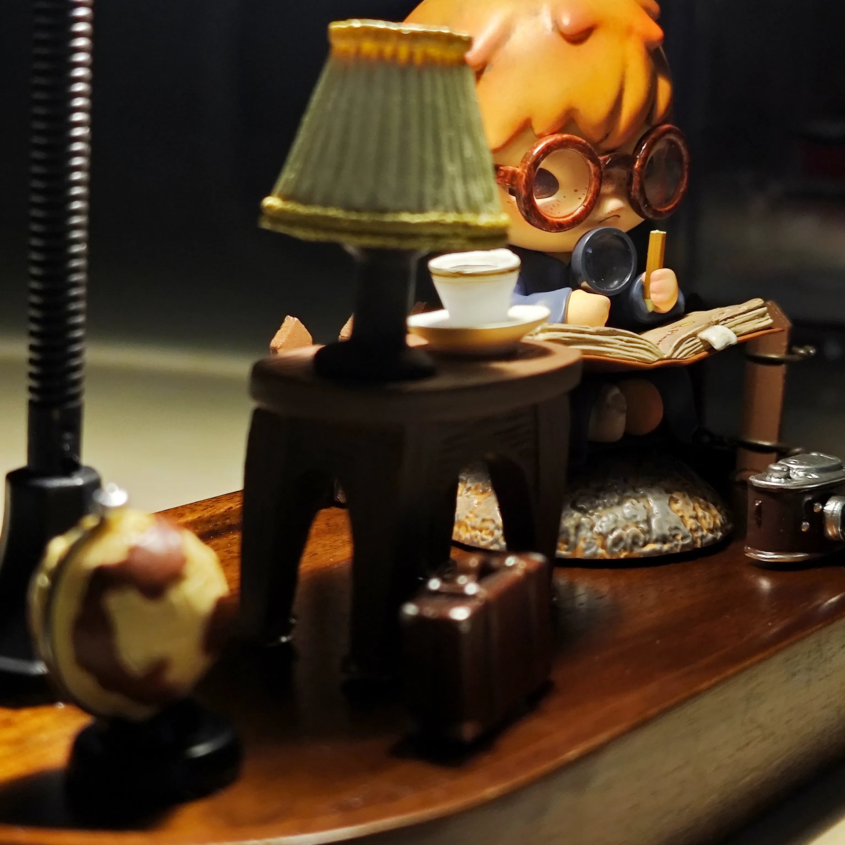 Side-angle view of Hirono the Geographer diorama showing detailed accessories including globe, suitcase, and camera with warm lamp illumination.