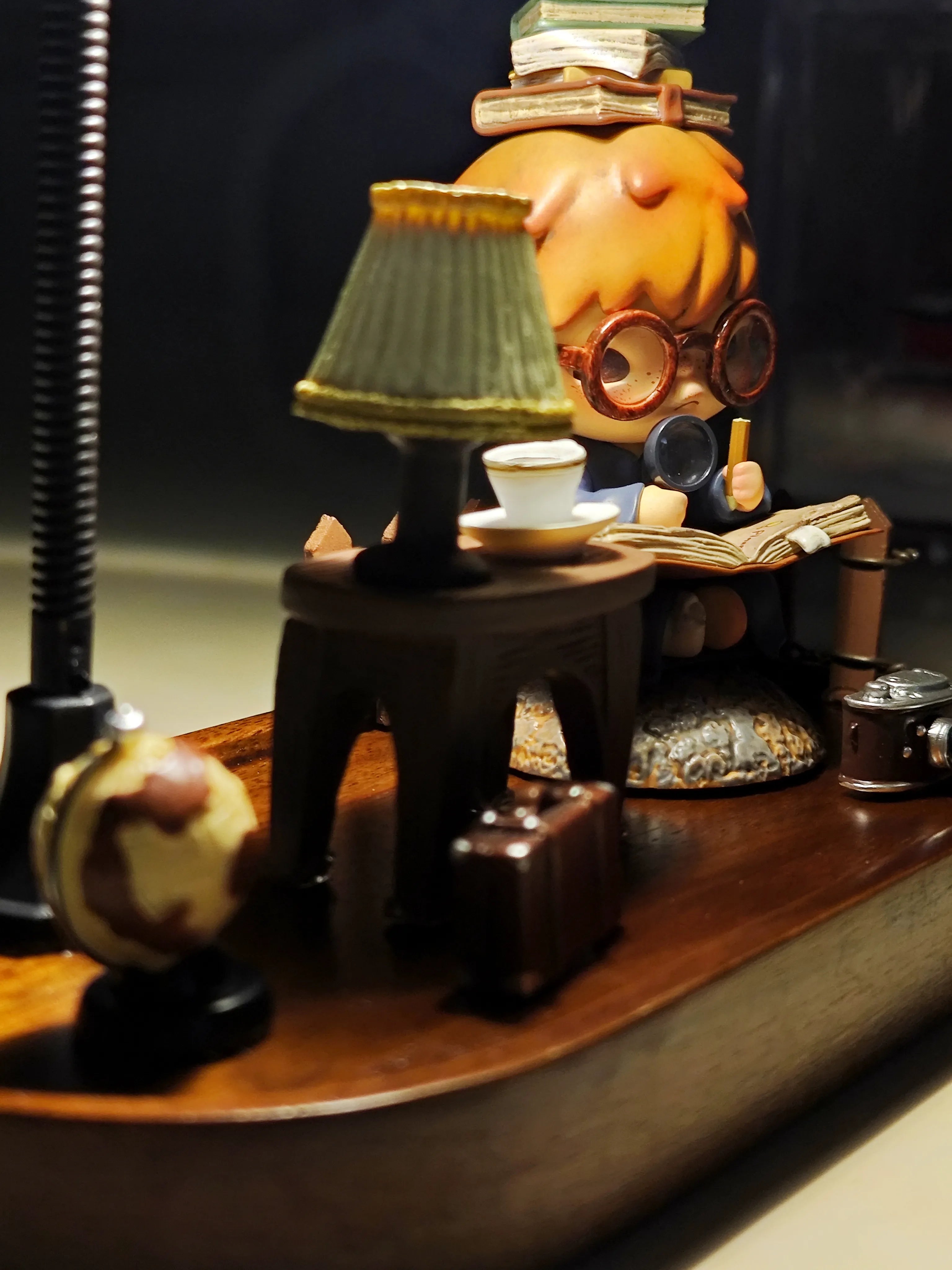 Side-angle view of Hirono the Geographer diorama showing detailed accessories including globe, suitcase, and camera with warm lamp illumination.