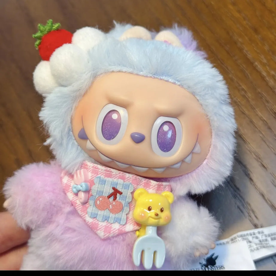 Labubu with a fluffy white head and pink body, wearing a bib with a cartoon character on a wooden surface.