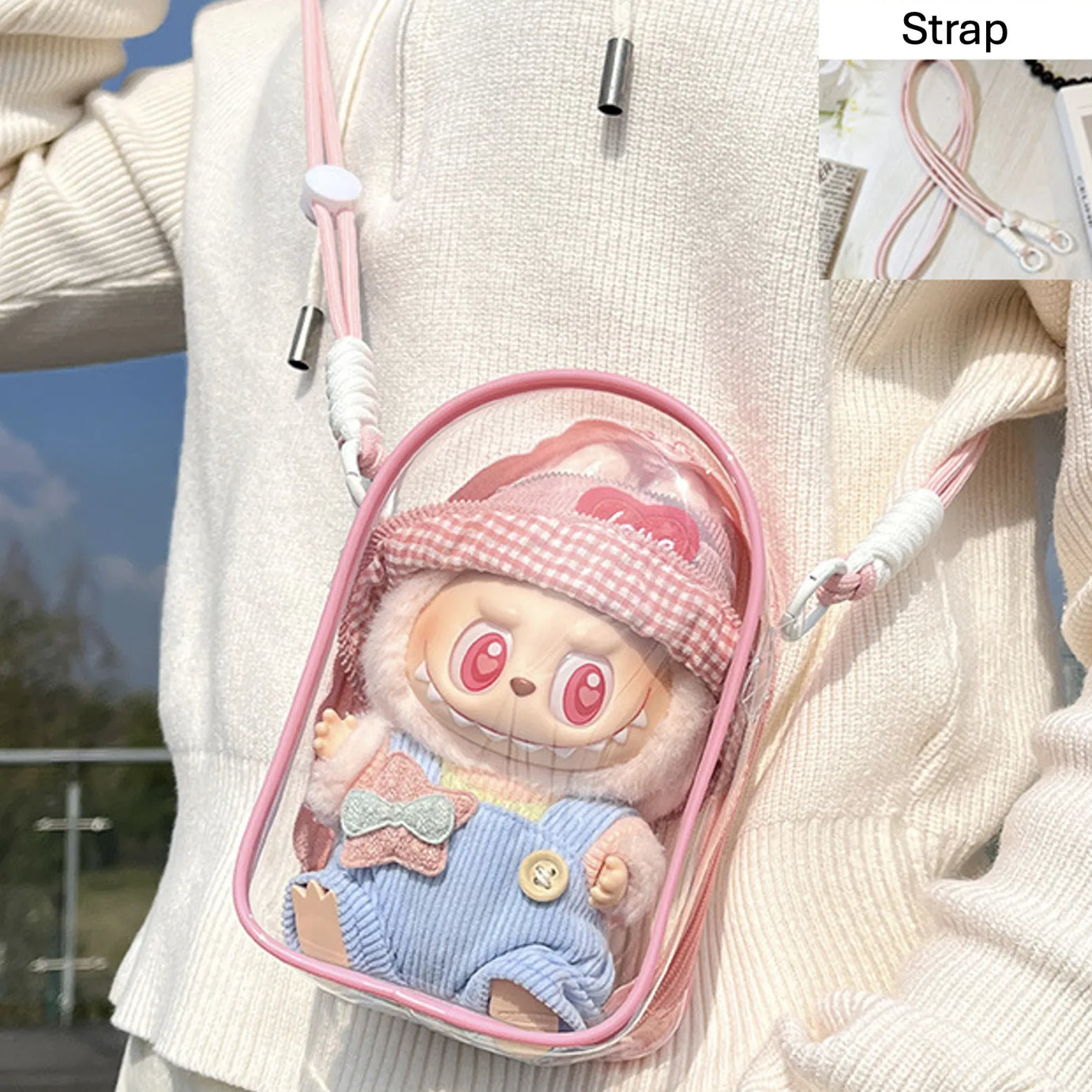 Transparent PVC display bag with a peach pink strap, showcasing a Labubu collectible figure dressed in pastel overalls and a gingham pink hat. The lightweight bag is compact and dustproof, perfect for collectors who want to showcase their small figures stylishly.