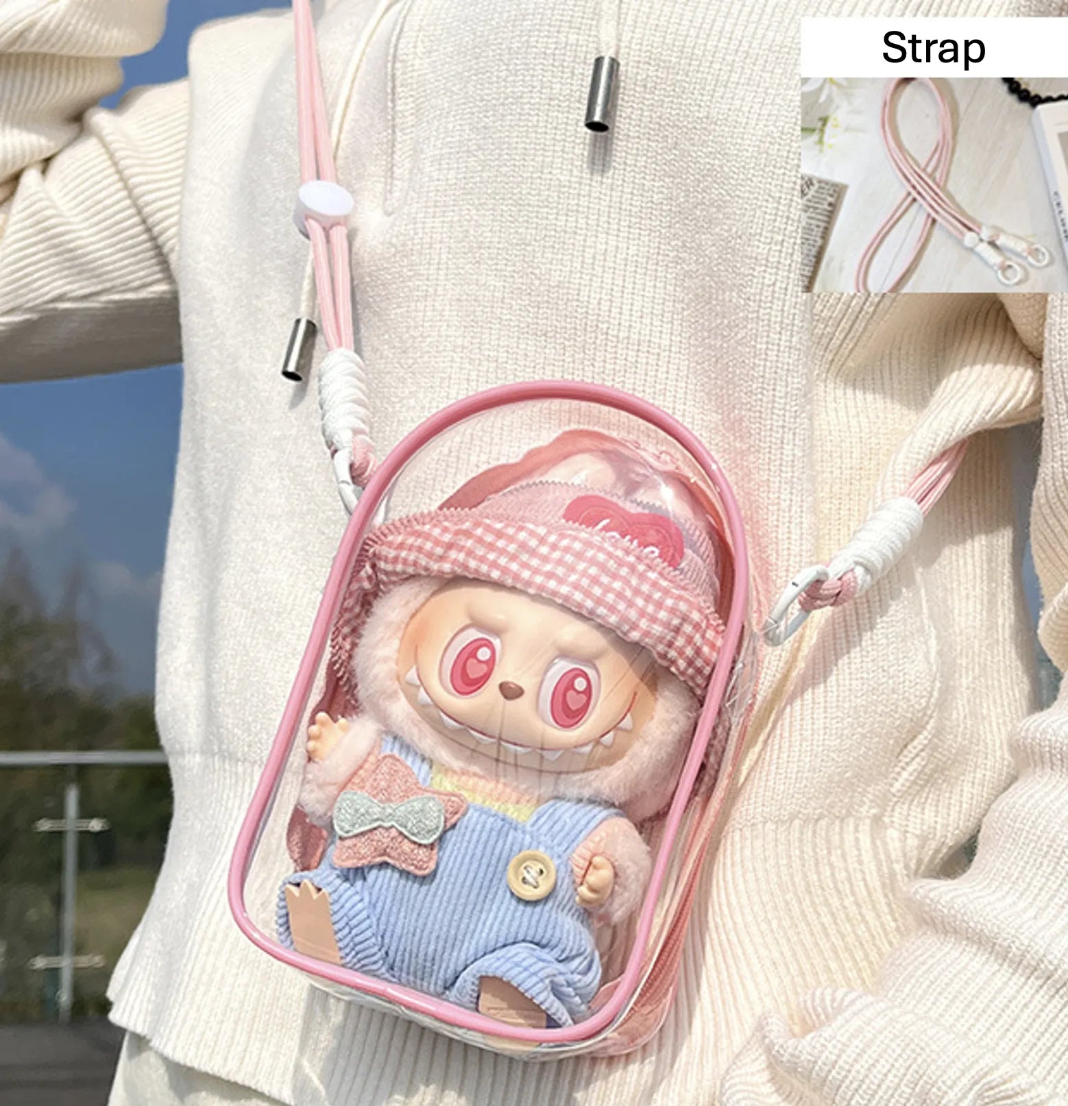 Transparent PVC display bag with a peach pink strap, showcasing a Labubu collectible figure dressed in pastel overalls and a gingham pink hat. The lightweight bag is compact and dustproof, perfect for collectors who want to showcase their small figures stylishly.